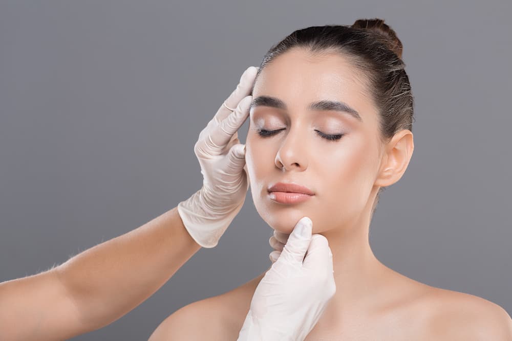 A woman with her eyes closed and hair in a bun against a solid grey background. A pair of hands in white medical gloves are gently cupping her face—one at the temple and one under the chin—as if performing an aesthetic assessment or marking for surgery.