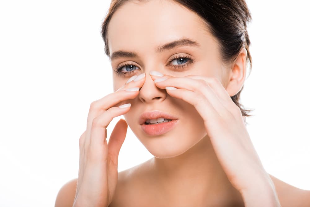 A young woman with dark hair and blue eyes looking directly at the camera against a plain white background. She is gently touching both sides of her nose with her fingers, suggesting a focus on the nasal structure or post-operative sensitivity.