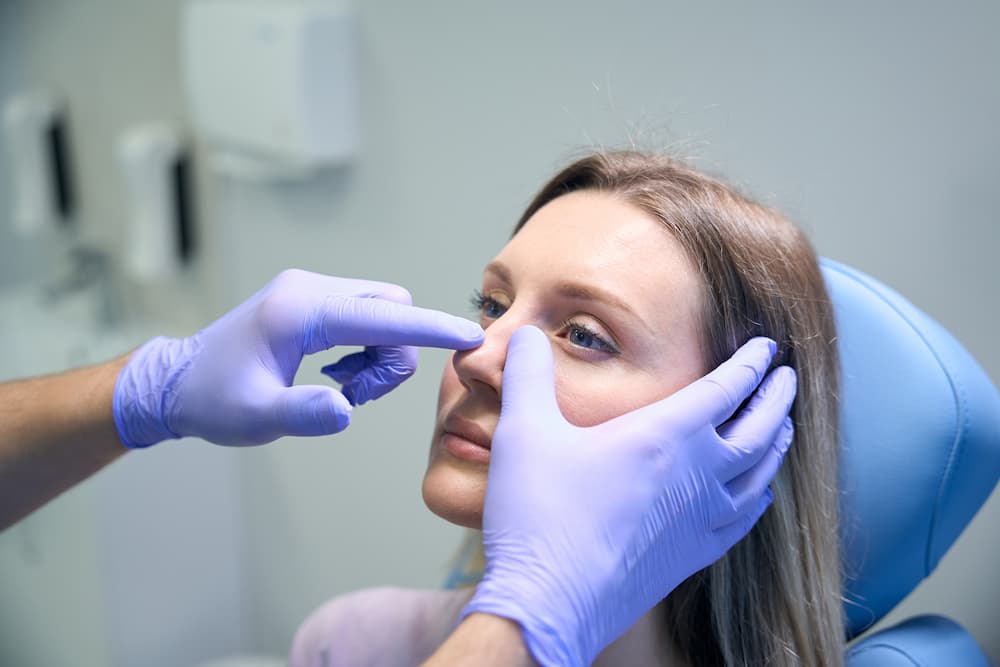 A close-up of a medical professional wearing purple nitrile gloves examining a blonde woman’s nose. The patient is sitting in a blue clinical chair, looking upward as the doctor assesses the nasal bridge and tip in a bright office setting
